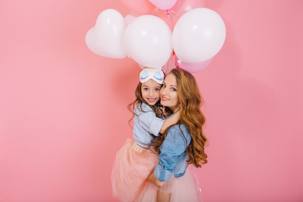 Retrato de niña linda del cumpleaños de pelo largo con globos blancos abrazando a su joven mamá rizada después del evento. Encantadora madre posando con bonita hija en fiesta aislada sobre fondo rosa