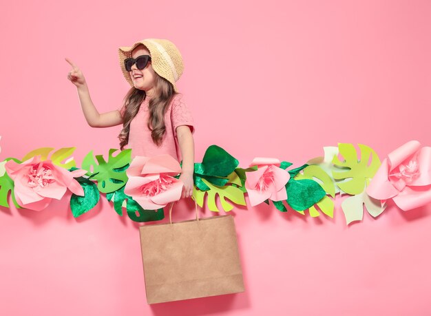 Retrato de niña linda con gafas y sombrero de verano, con bolsa de compras en mano sobre fondo rosa con flores de papel, lugar para texto, concepto de publicidad de verano