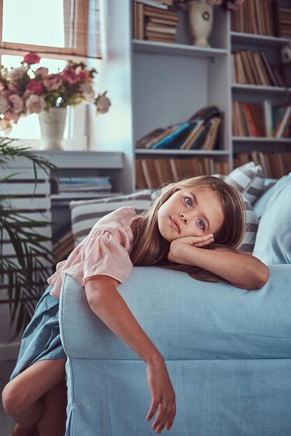 Retrato de una niña linda con el pelo largo y castaño y una mirada penetrante, mirando una cámara, tendida en un sofá solo en casa.