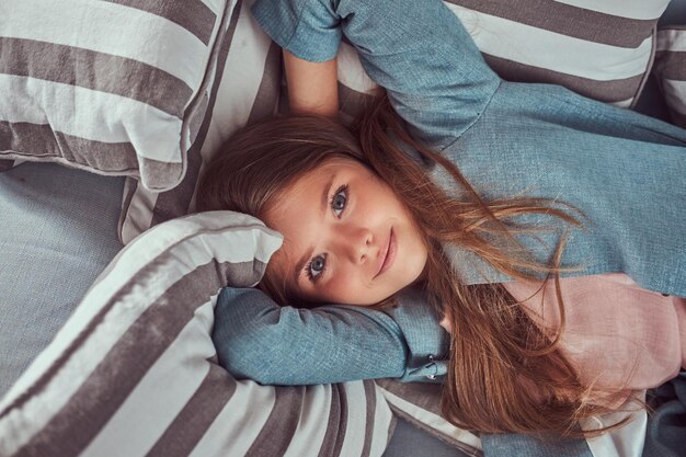 Foto gratuita retrato de una niña linda con el pelo largo y castaño, mirada penetrante y sonrisa encantadora, mirando una cámara, recostada en un sofá en casa sola.