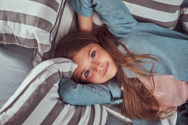 Retrato de una niña linda con el pelo largo y castaño, mirada penetrante y sonrisa encantadora, mirando una cámara, recostada en un sofá en casa sola.