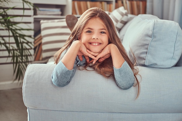 Retrato de una niña linda con el pelo largo y castaño, mirada penetrante y sonrisa encantadora, mirando una cámara, recostada en un sofá en casa sola.