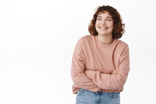 Foto gratuita retrato de niña linda ríe y sonríe, de pie relajado en pose casual, brazos cruzados sobre el pecho como si sintiera frío, mirando a un lado en la esquina superior izquierda, de pie sobre blanco.