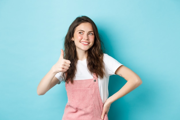 Retrato de niña linda sonriente con peinado rizado, mostrando el pulgar hacia arriba y mirando pensativo en el banner de la esquina superior izquierda, alabando la venta de la tienda, de pie contra el fondo azul.