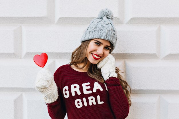 Retrato de niña en marsala sweater y gorro de punto en la pared gris exterior. Lleva guantes blancos, sostiene una piruleta de corazón rojo y sonríe.