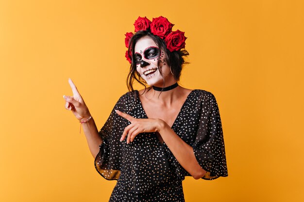 Retrato de niña mexicana positiva sobre fondo naranja con espacio para texto. Mujer con máscara de calavera sonríe tiernamente y señala con los dedos hacia arriba.