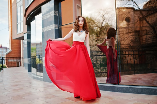 Retrato de niña de moda en el vestido de noche rojo que plantea la ventana de espejo de fondo del edificio moderno