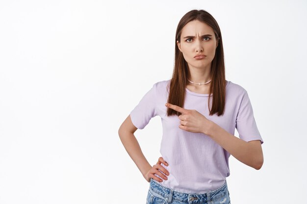 Retrato de niña molesta apuntando hacia la izquierda, haciendo muecas ante algo injusto, quejarse, parece decepcionado y molesto, de pie sobre blanco