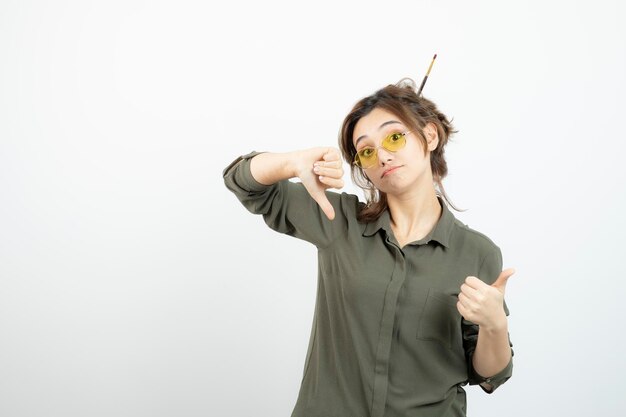 Retrato de niña con moño desordenado en gafas de pie y dando pulgares hacia arriba. foto de alta calidad