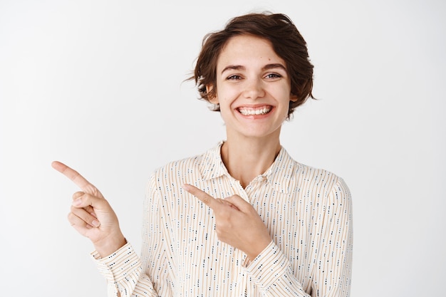 Retrato de niña morena alegre en blusa apuntando con el dedo hacia la izquierda, sonriendo y mostrando compañía, demostrar promoción, de pie sobre una pared blanca