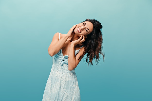 Retrato de niña morena alegre en vestido sobre fondo azul. Linda chica adulta juega con su pelo oscuro ondulado corto y mira a la cámara.