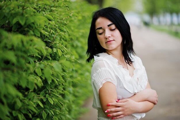 Retrato de niña morena en blusa blanca de mujer contra arbustos verdes de primavera