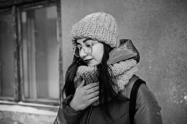 Retrato de niña morena en bufanda gris y gafas de sombrero en clima frío con sol contra la pared naranja de la casa antigua