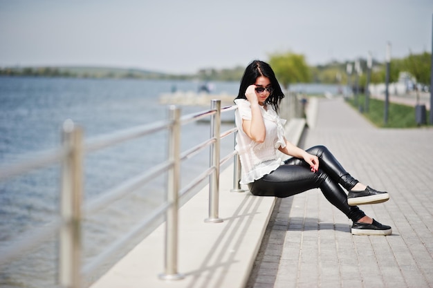 Retrato de niña morena en pantalones de cuero de mujer y gafas de sol de blusa blanca contra barandillas de hierro en la playa