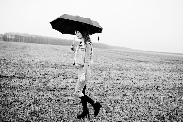 Retrato de niña morena rizada en chaqueta de jeans con paraguas negro en el campo