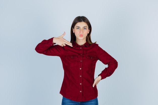 Retrato de niña mostrando gesto de pistola, haciendo pucheros con los labios en camisa burdeos y mirando confiado