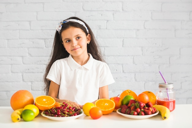 Retrato de una niña con muchas frutas en el escritorio blanco contra la pared