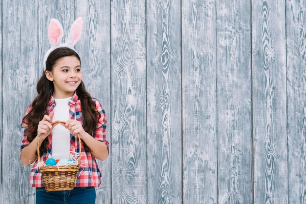 Retrato de una niña con orejas de conejo con canasta de huevos de Pascua mirando lejos