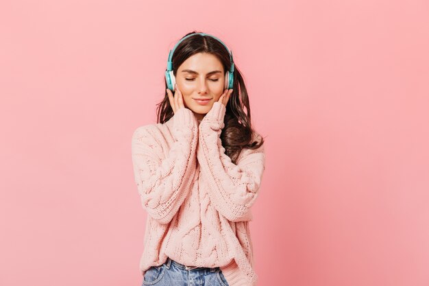 Retrato de niña pacificada escuchando agradable melodía en auriculares. Señora en suéter lindo sonriendo con los ojos cerrados sobre fondo rosa.