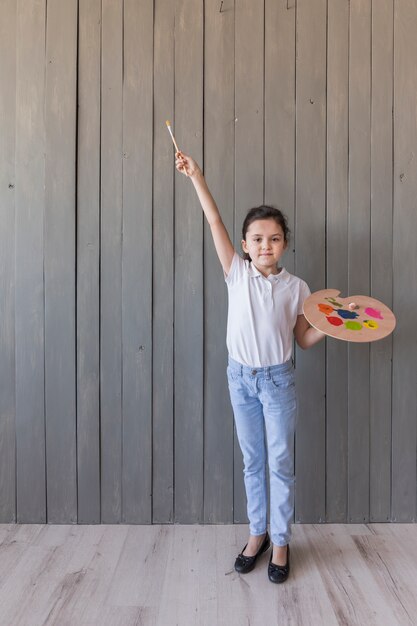 Retrato de una niña con paleta pintada y pincel de pie contra un tablón de madera gris