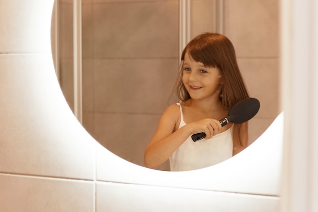Retrato de niña peinándose en el baño, haciendo procedimientos de belleza matutinos por su cuenta mientras está de pie frente al espejo, vistiendo ropa de casa.