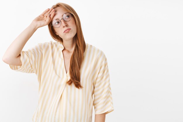 Retrato de niña pelirroja triste sin emoción cansada con pecas en gafas y blusa amarilla azotando sudor en la frente mirando a la derecha con mirada agotada