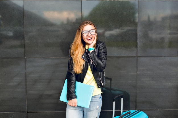 Retrato de niña con pelo largo con gafas está de pie afuera sobre fondo negro. Viste chaqueta negra con jeans y sostiene una computadora portátil. Ella sonríe a la cámara.