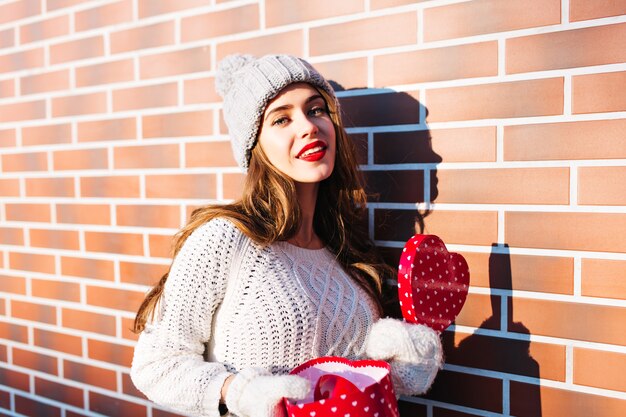 Retrato de niña con pelo largo en gorro de punto, suéter cálido y guantes en la pared exterior. Ella sostiene el corazón de caja abierta en las manos, sonriendo.