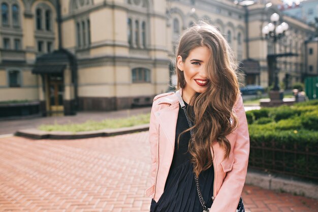 Retrato de niña con pelo largo y rizado y labios rojos Lleva vestido negro, chaqueta rosa. El cabello cubre la mitad de su rostro y ella está sonriendo.