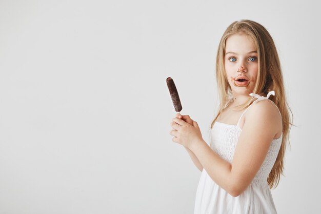 Retrato de niña pequeña graciosa guapa con el pelo largo rubio con expresión de la cara de sorpresa, estar sucio después de comer helado.