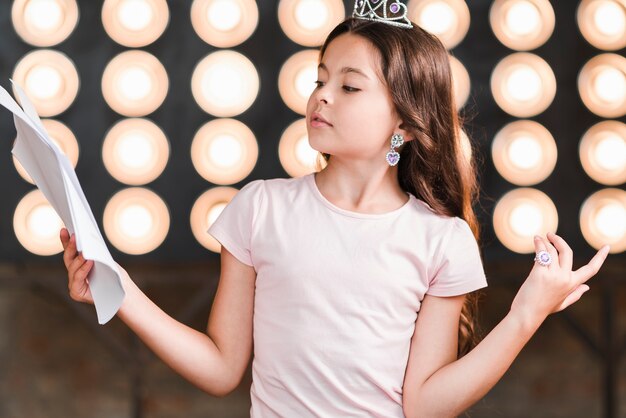 Retrato de niña de pie contra la luz del escenario mirando guiones encogiéndose de hombros