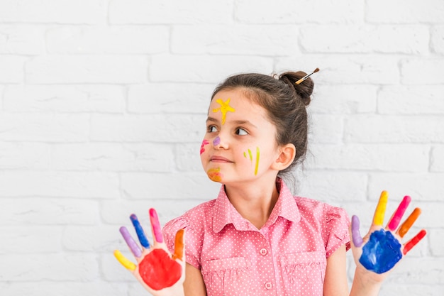 Retrato de una niña de pie contra la pared blanca que muestra las manos pintadas de colores mirando a otro lado