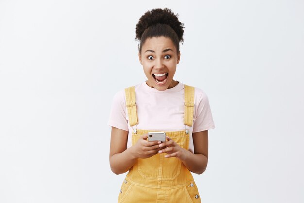 Retrato de niña de piel oscura guapa sorprendida y asombrada con un mono amarillo, gritando y sosteniendo el teléfono inteligente