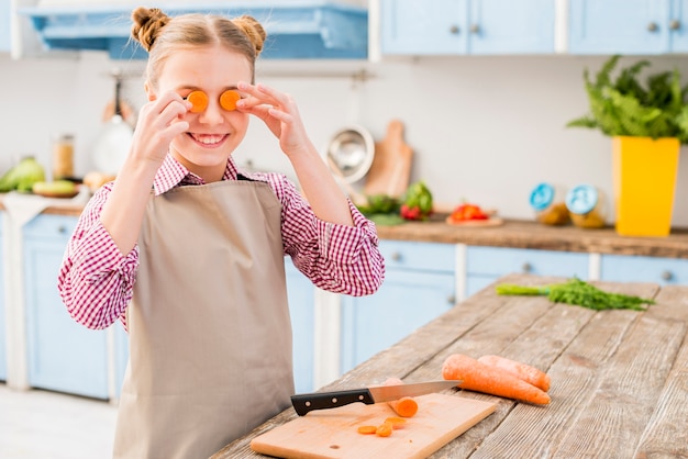 Retrato de una niña que cubre sus ojos con una rodaja de zanahoria en la cocina