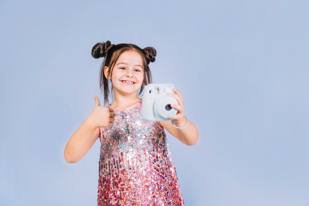 Retrato de una niña que sostiene la cámara instantánea que muestra el pulgar hacia arriba de la muestra contra el fondo azul