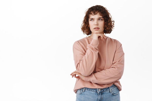 Retrato de niña rizada pensativa, estudiante haciendo suposiciones, mirada seria y pensando, mirando a la esquina superior izquierda enfocada, reflexionando sobre la decisión, de pie en blanco