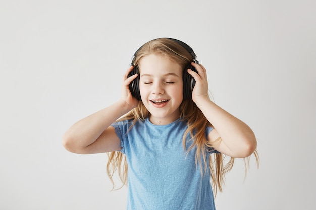 Retrato de niña rubia en camisa azul jugando con grandes auriculares inalámbricos, escuchando música, cantando canciones y bailando con los ojos cerrados mientras nadie en casa.