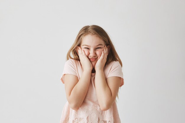 Retrato de niña rubia divertida en vestido rosa, apretando la cara con las manos, haciendo caras tontas para evitar que la madre le tome una buena foto.