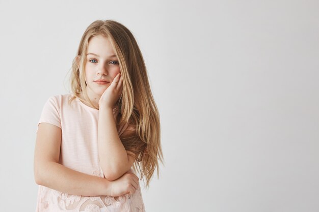 Retrato de niña rubia linda en camiseta rosa sosteniendo la cabeza con la mano, estar cansado y aburrido durante las clases escolares.