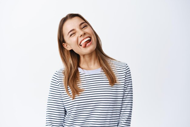 Retrato de niña rubia positiva divirtiéndose, sonriendo dientes blancos y mostrando la lengua, guiñando un ojo sin preocupaciones, sentirse feliz, de pie sobre blanco