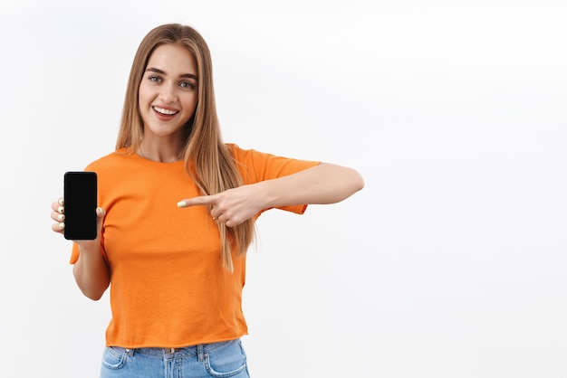 Retrato de niña rubia sonriente alegre y feliz mediante teléfono móvil, señalando con el dedo a la pantalla del teléfono inteligente, mostrando el sitio de compras