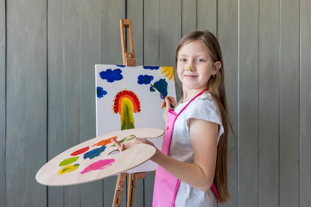 Retrato de una niña rubia sonriente que sostiene la paleta en la pintura de la mano en la base con el pincel
