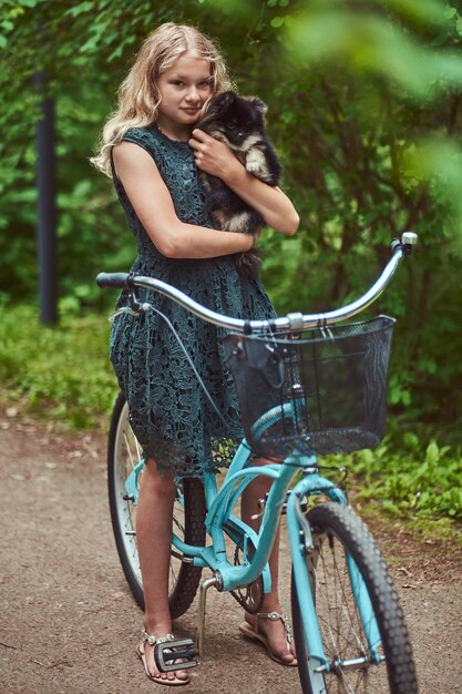 Retrato de una niña rubia con un vestido informal, tiene un lindo perro spitz. Andar en bicicleta en un parque.