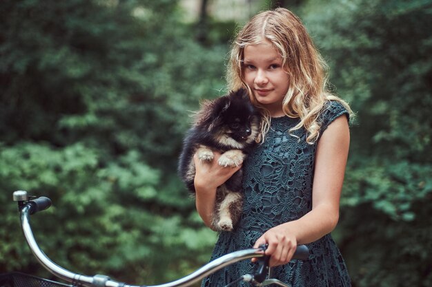 Retrato de una niña rubia con un vestido informal, tiene un lindo perro spitz. Andar en bicicleta en un parque.