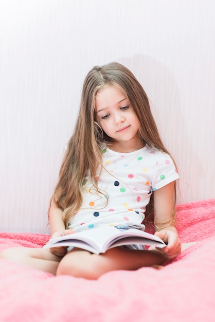 Retrato de una niña sentada en el libro de lectura de cama de color rosa suave