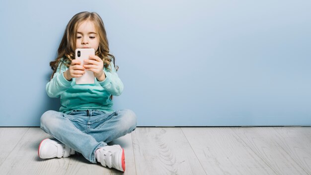 Retrato de una niña sentada en el piso de madera dura mirando smartphone