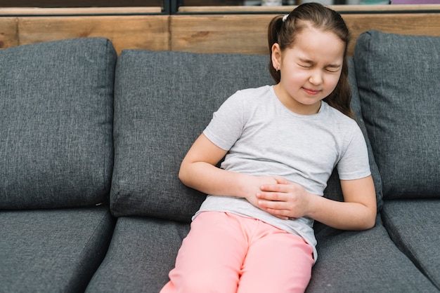 Retrato de una niña sentada en un sofá gris que tiene dolor severo en el estómago