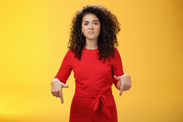 Retrato de niña sombría con cara triste frunciendo los labios frunciendo el ceño decepcionada y apuntando hacia abajo con arrepentimiento y expresión insatisfecha, perdiendo la oportunidad sintiéndose molesto por el fondo amarillo.