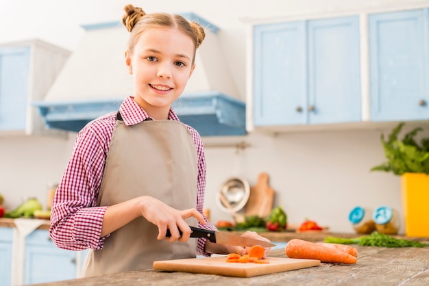 Retrato de niña sonriente cortando la zanahoria con un cuchillo en la mesa mirando a la cámara