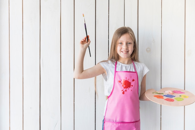 Foto gratuita retrato de una niña sonriente en delantal con pincel y paleta de madera en la mano contra la pared de tablones de madera